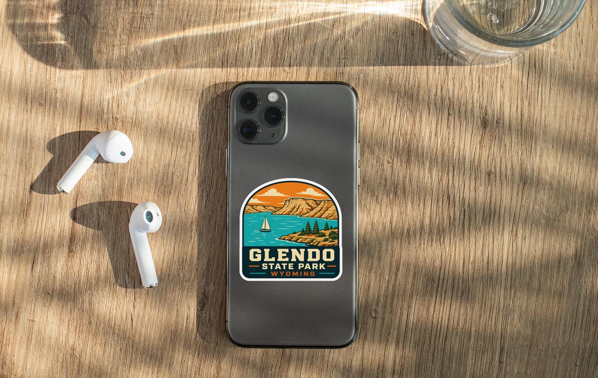 a smartphone with a sticker on the back, placed on a wooden surface alongside a pair of white earbuds and a glass of water.