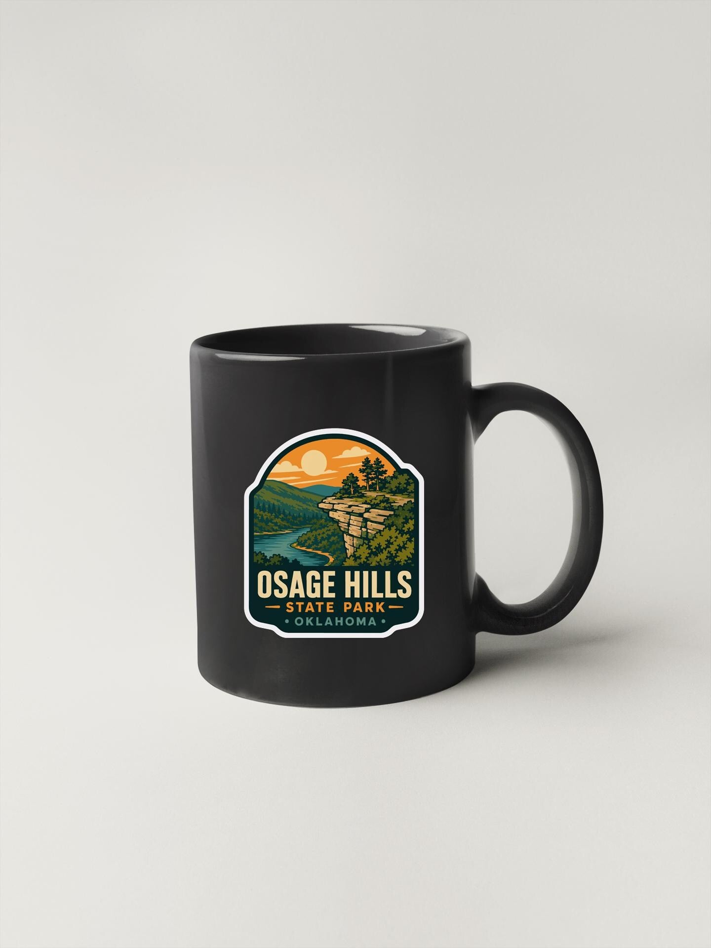A black coffee mug with a sticker depicting a scenic landscape, including a cliff, river, and sunset.