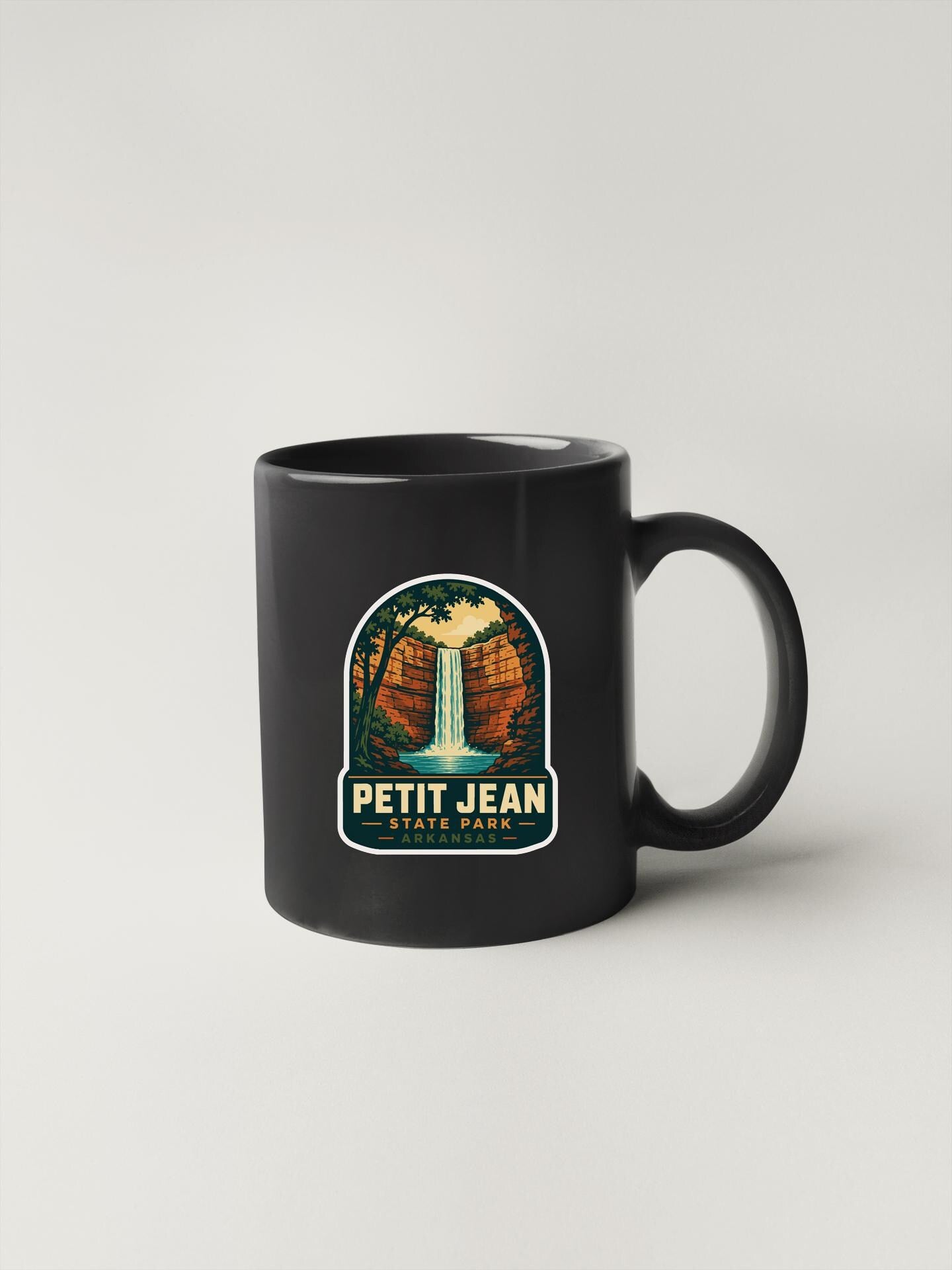 A black coffee mug with a sticker depicting a waterfall and the text "PETIT JEAN STATE PARK".