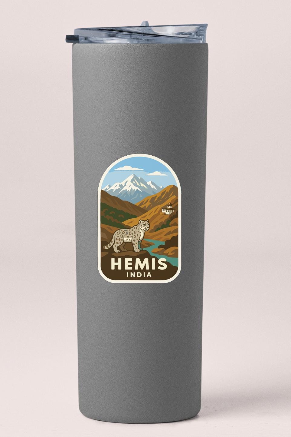 A gray tumbler with a sticker depicting a mountain landscape and a snow-capped peak, along with the text "HEMIS INDIA".