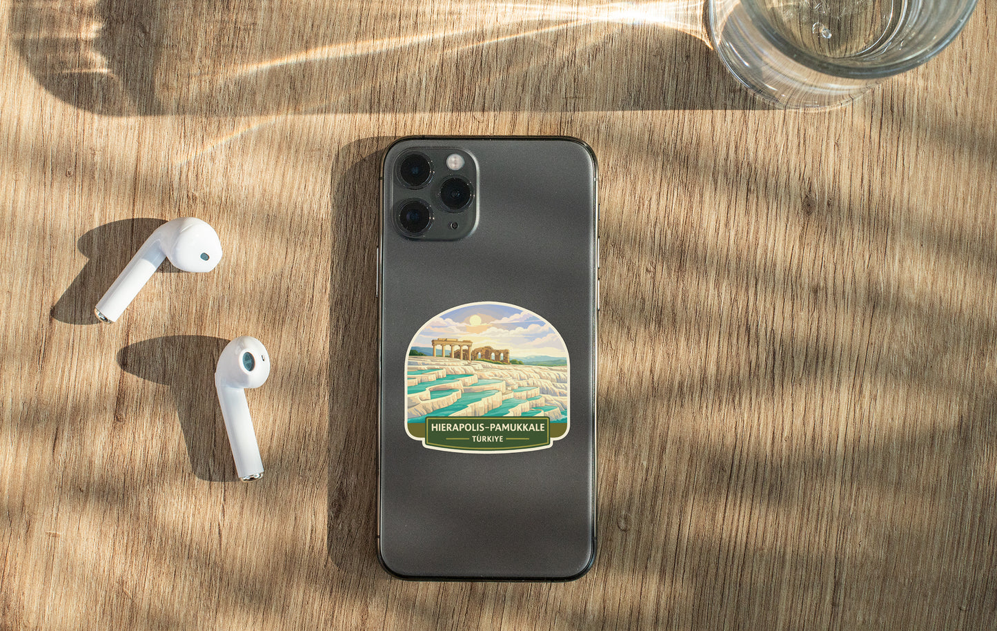 a smartphone with a sticker on the back, a pair of white earbuds, and a glass of water on a wooden surface.