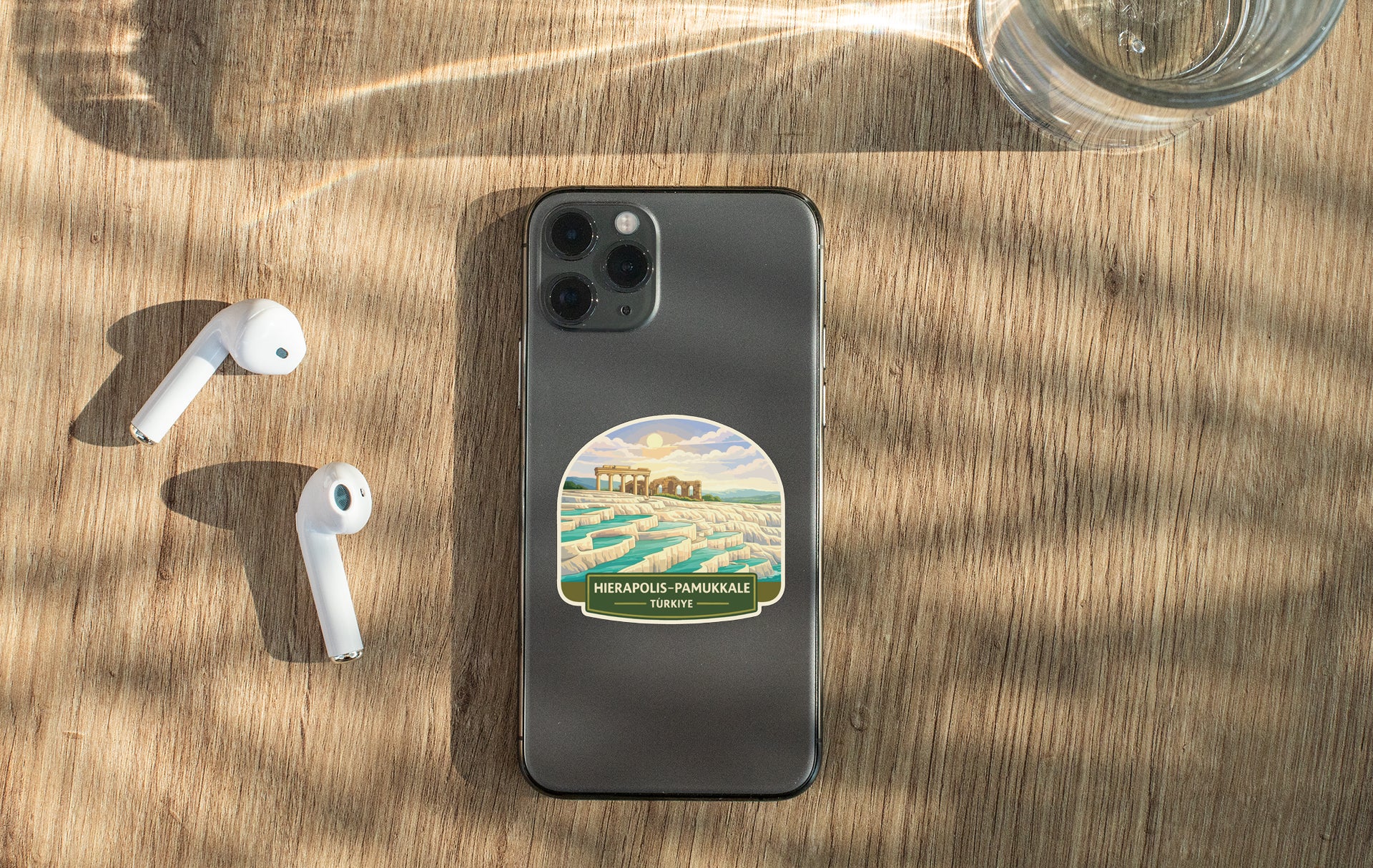 a smartphone with a sticker on the back, a pair of white earbuds, and a glass of water on a wooden surface.