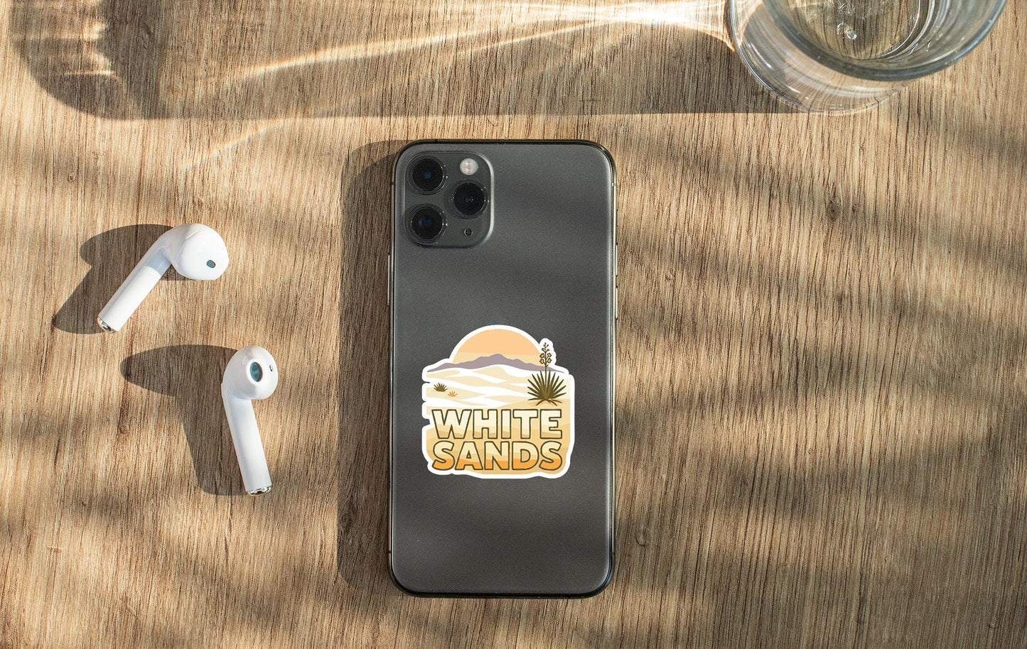 a smartphone with a sticker on the back, placed on a wooden surface alongside a pair of white earbuds and a glass of water.