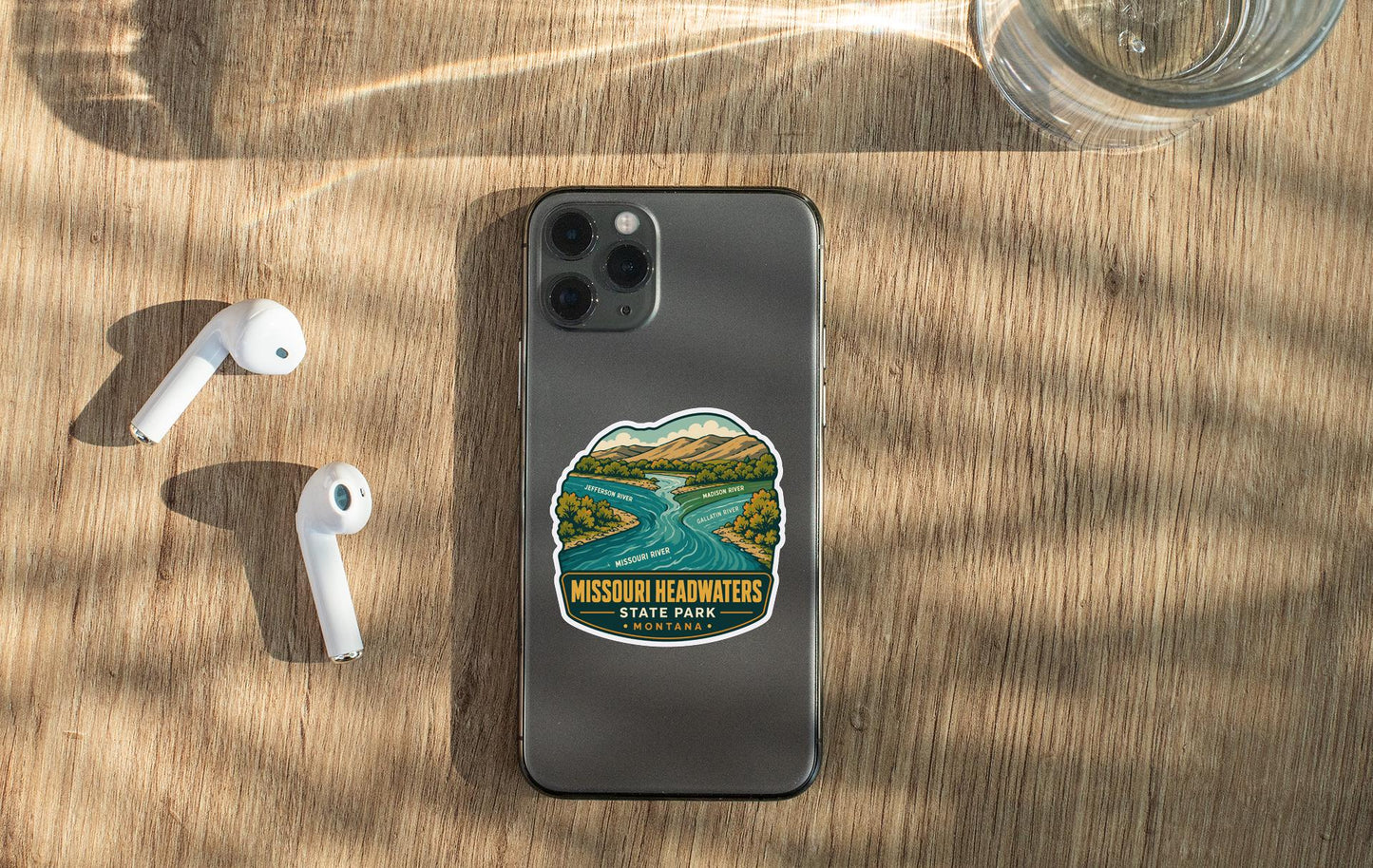 a smartphone with a Missouri Reachwaters State Park sticker on it, placed on a wooden surface alongside a pair of white earbuds and a glass of water.