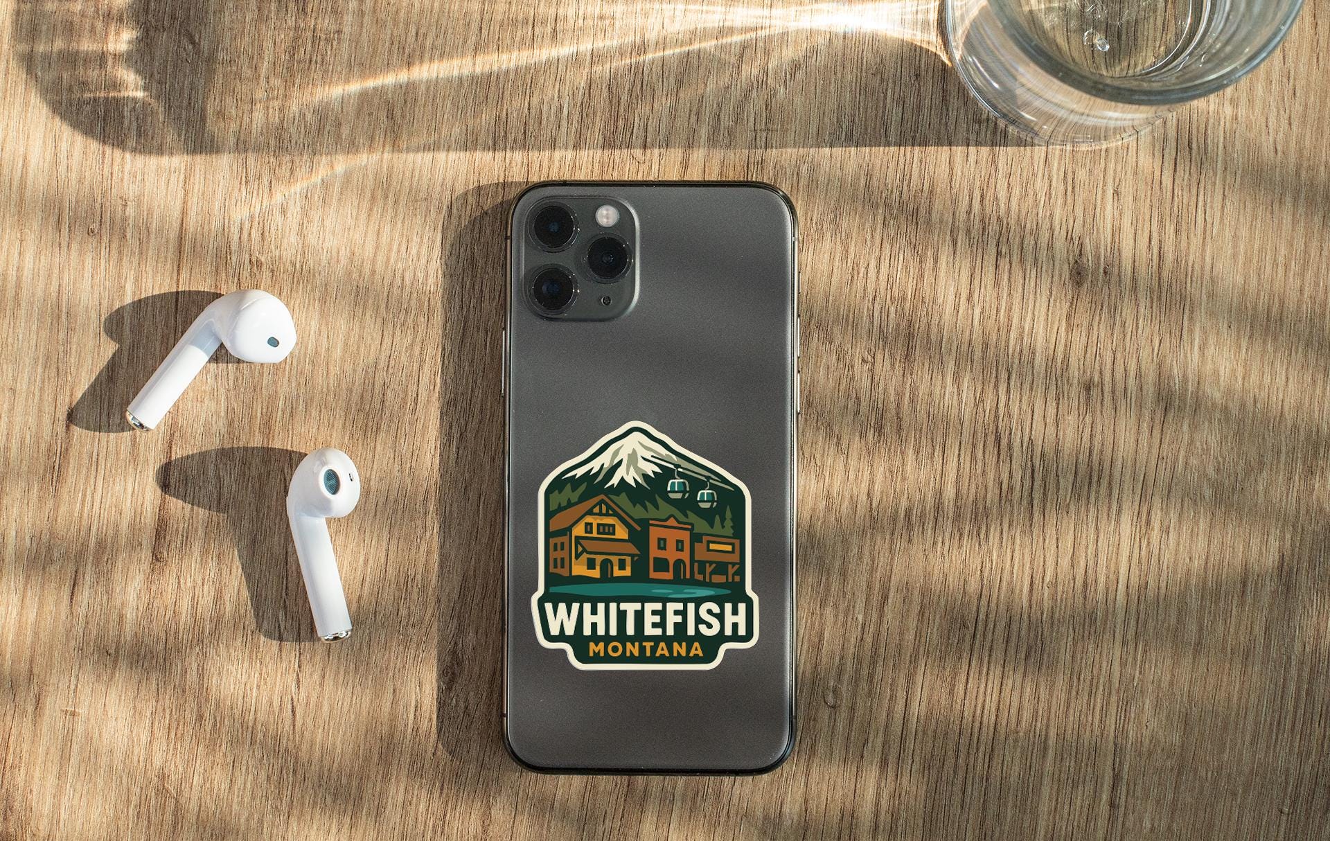a smartphone with a sticker on the back, placed on a wooden surface alongside a pair of white earbuds and a glass of water.