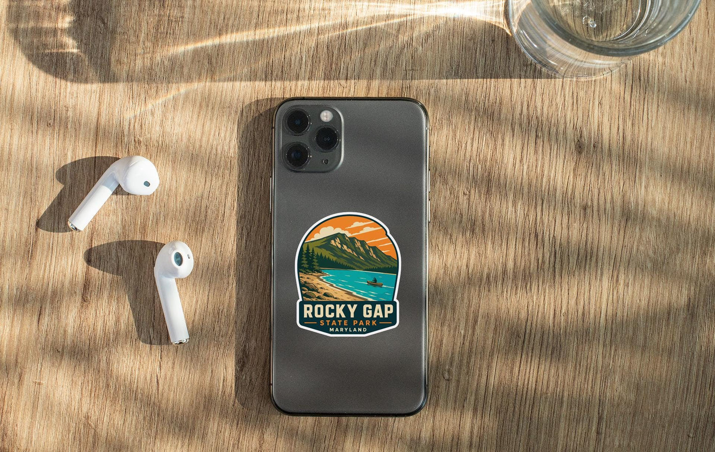 a smartphone with a sticker on the back, placed on a wooden surface alongside a pair of white earbuds and a glass of water.