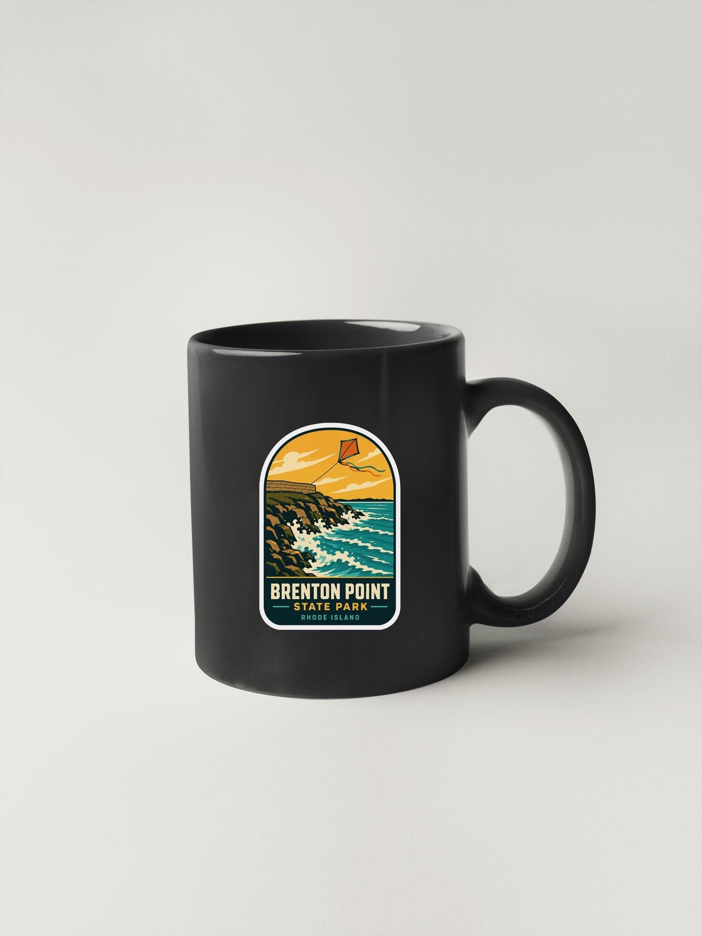 A black coffee mug with a graphic design depicting a coastal scene, including a beach, ocean, and mountains.