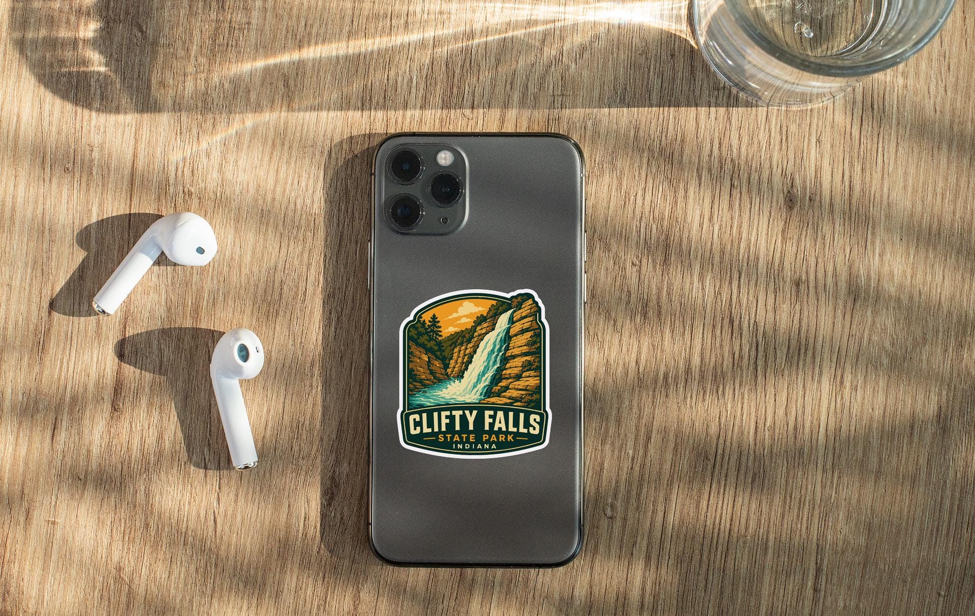 a smartphone with a sticker of Clifty Falls State Park on its back, along with a pair of white earbuds and a glass of water on a wooden surface.