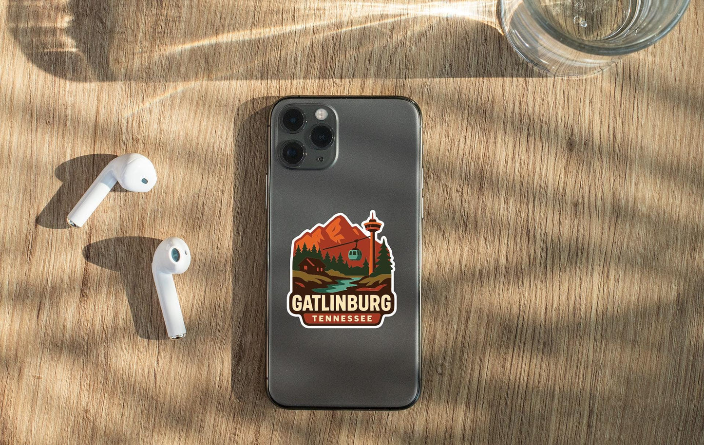 a smartphone with a sticker of a mountain landscape on it, placed on a wooden surface alongside a pair of white earbuds and a glass of water.
