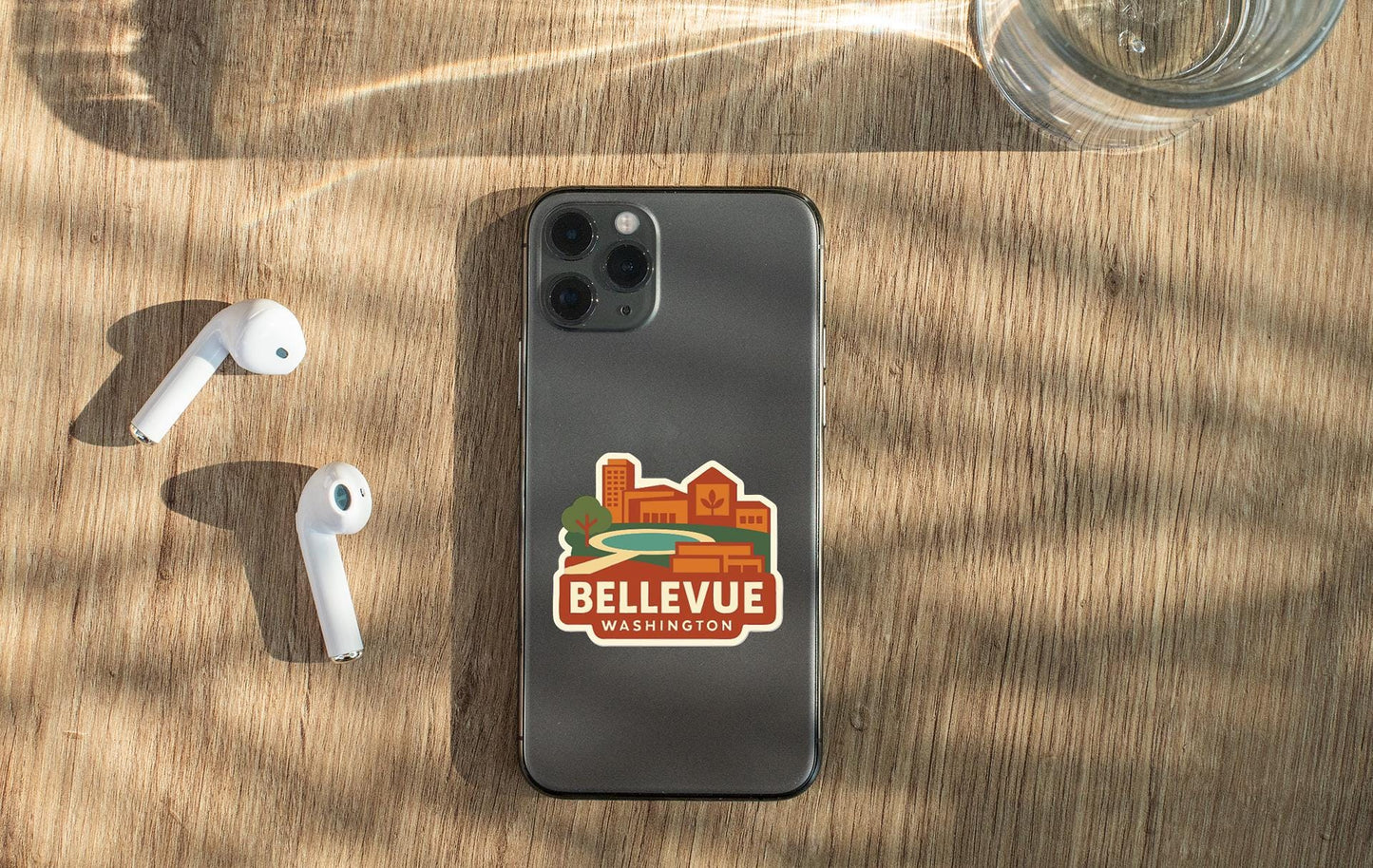 a smartphone with a sticker on the back, placed on a wooden surface alongside a pair of white earbuds and a glass of water.