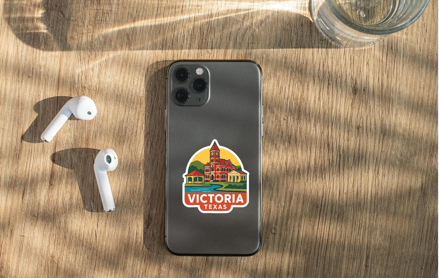 a smartphone with a Victoria, Texas sticker on the back, placed on a wooden surface alongside a pair of white earbuds and a glass of water.