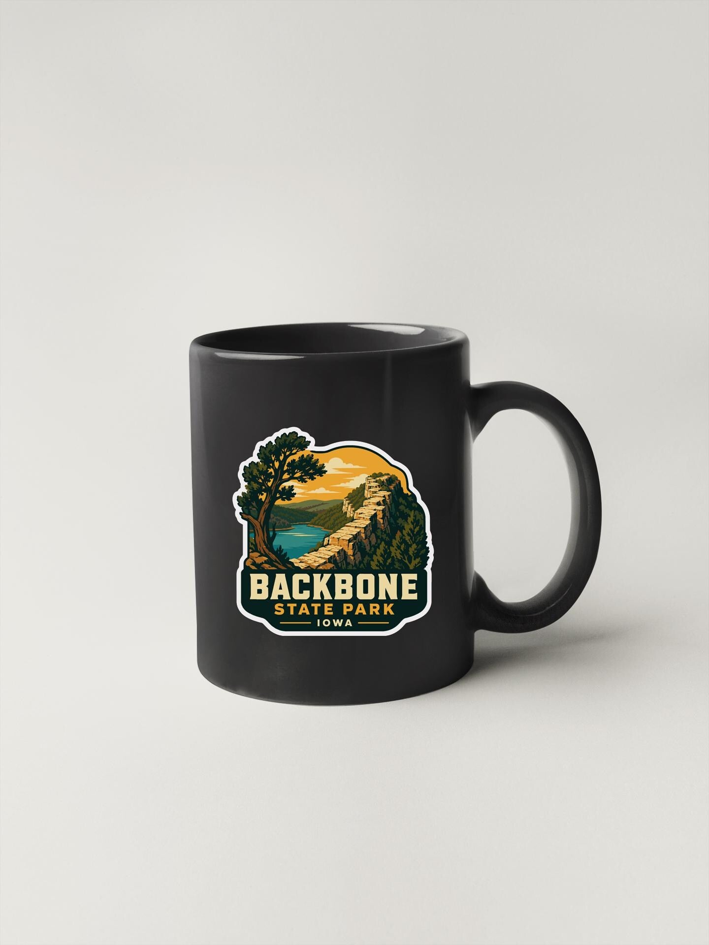 A black coffee mug with a sticker depicting a scenic landscape, including a mountain, lake, and trees, along with the text "BACKBONE STATE PARK, ID".