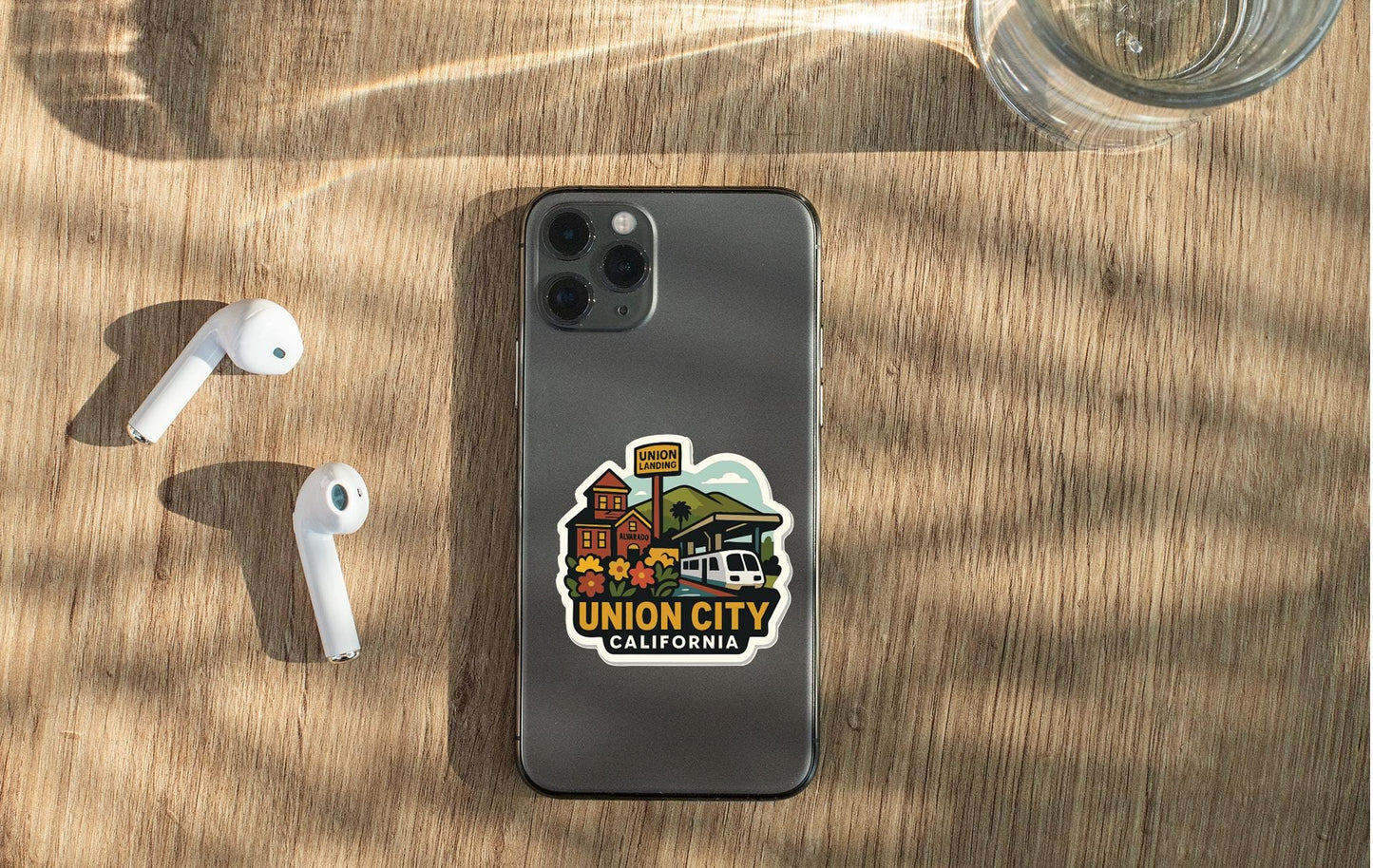 a smartphone with a sticker on it, placed on a wooden surface alongside a pair of white earbuds and a glass of water.