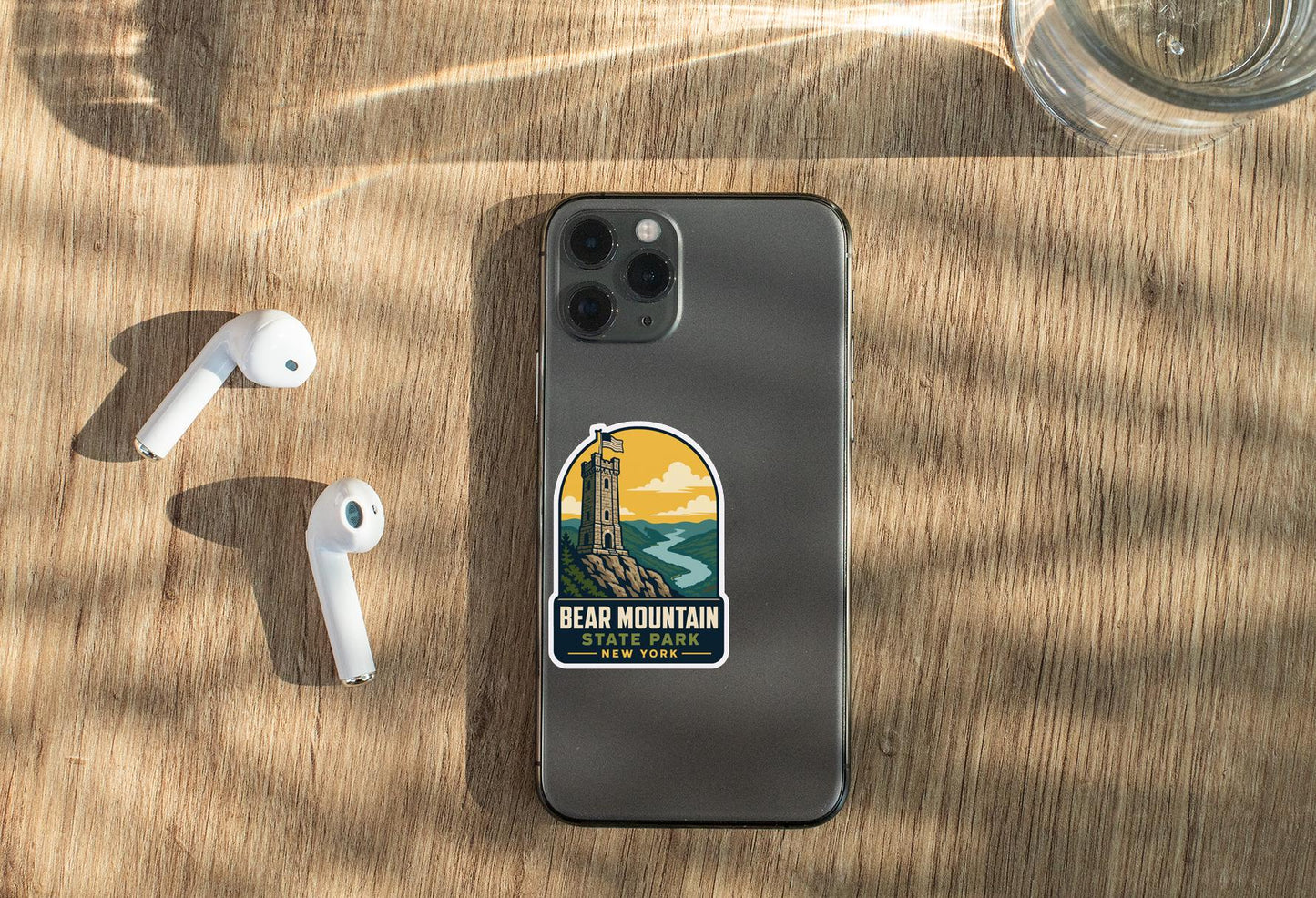 a smartphone with a sticker on the back, placed on a wooden surface alongside a pair of white earbuds and a glass of water.