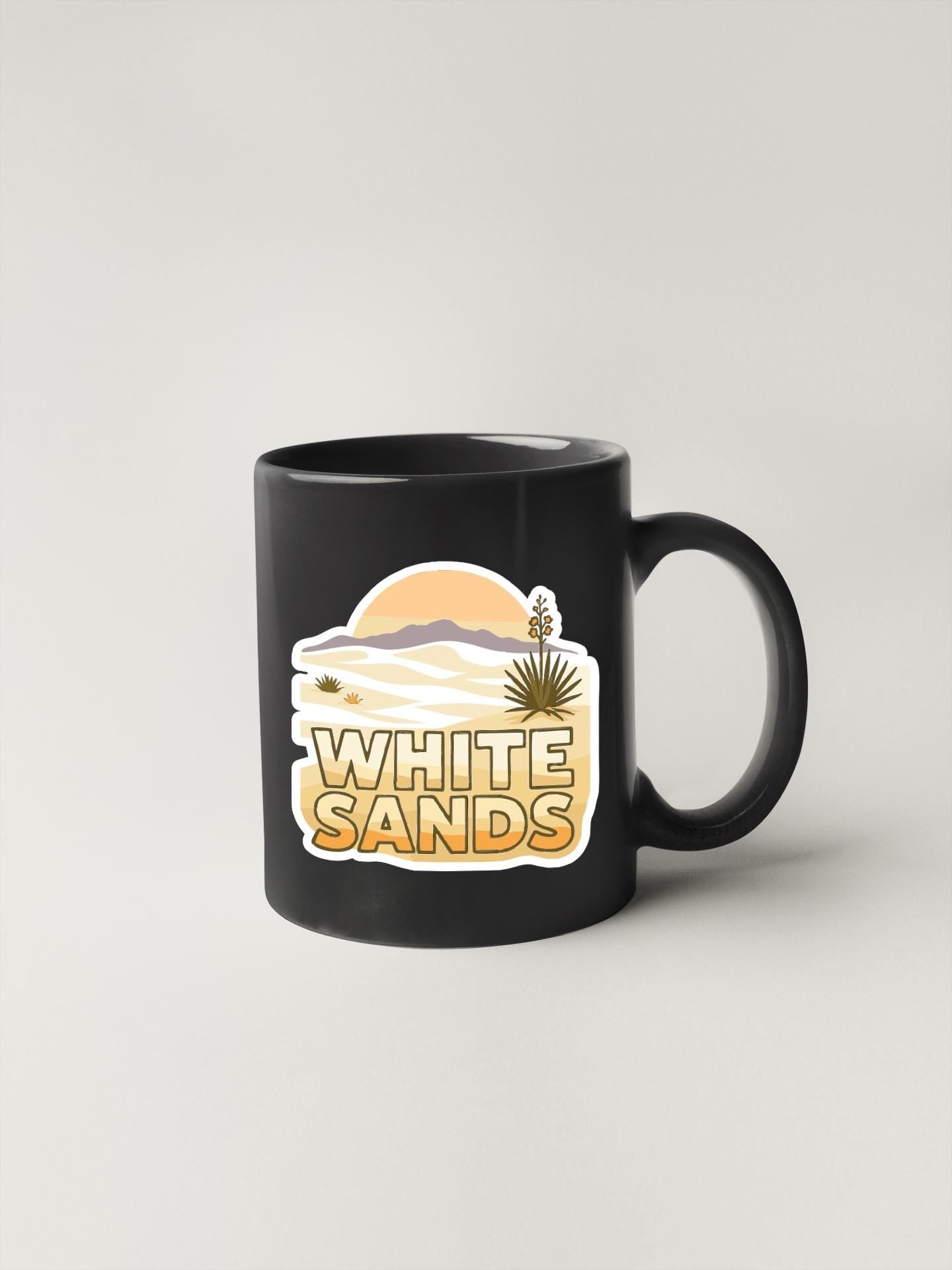 A black coffee mug with a graphic design featuring a desert landscape and the text "WHITE SAND".