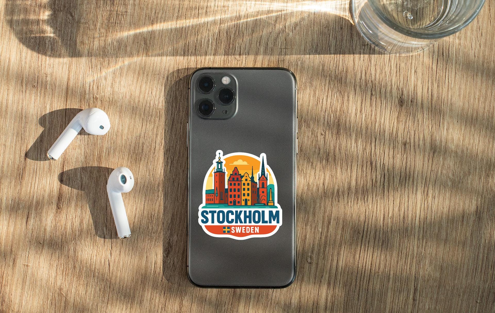 a smartphone with a sticker of Stockholm, Sweden, placed on a wooden surface alongside a pair of white earbuds and a glass of water.