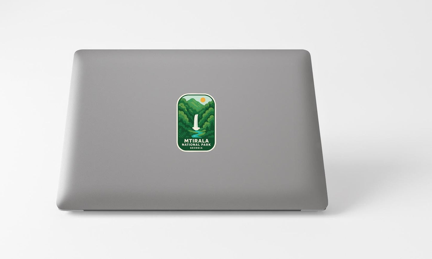 A sticker with a mountain landscape and the text "MTIRALA NATIONAL PARK" is placed on the back of a grey laptop computer.