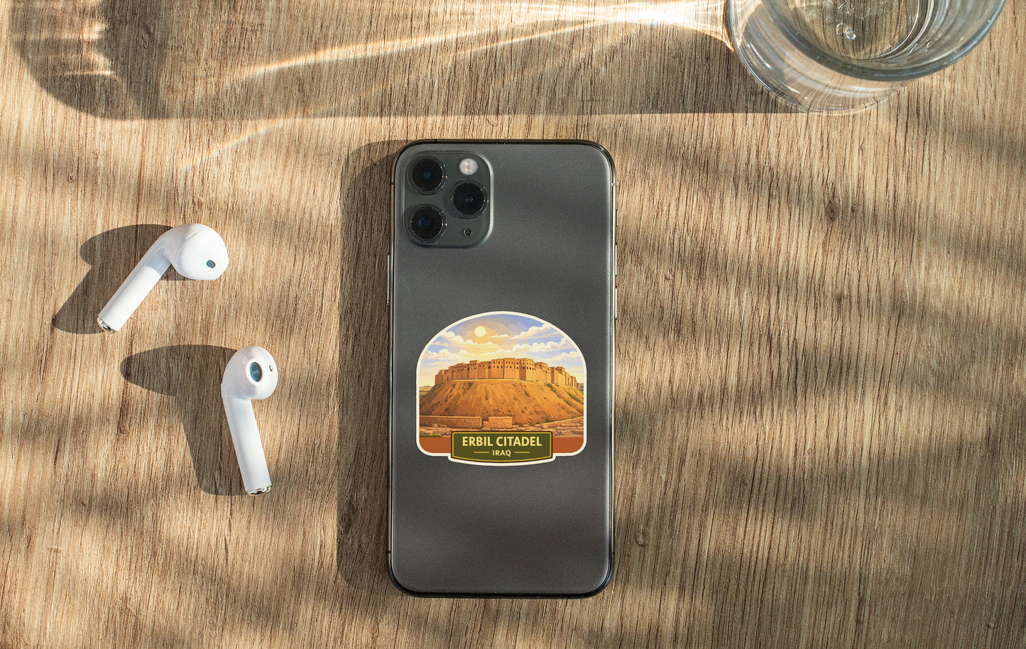 a smartphone with a sticker of a desert landscape on its back, along with a pair of white earbuds and a glass of water on a wooden surface.