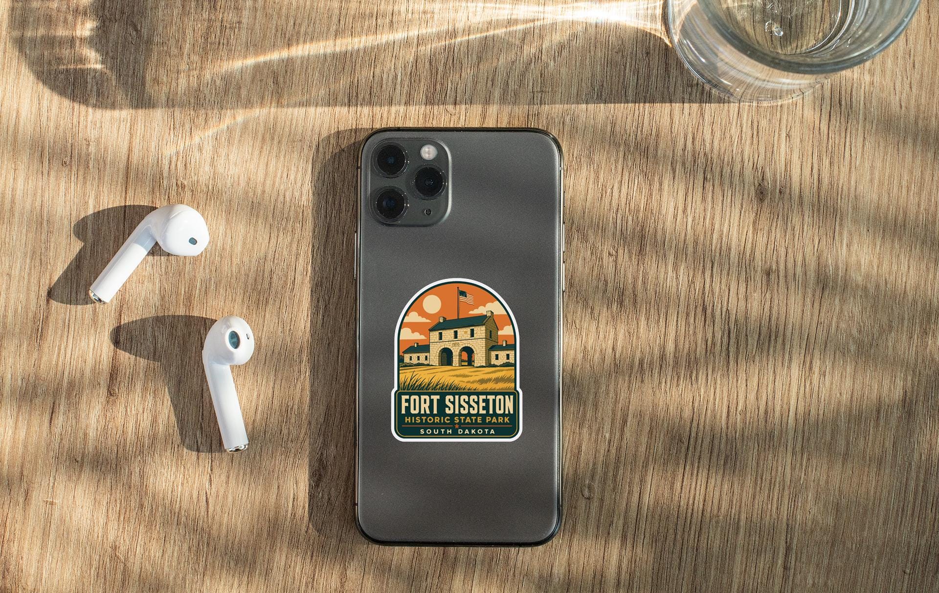 a smartphone with a sticker on it, placed on a wooden surface alongside a pair of white earbuds and a glass of water.
