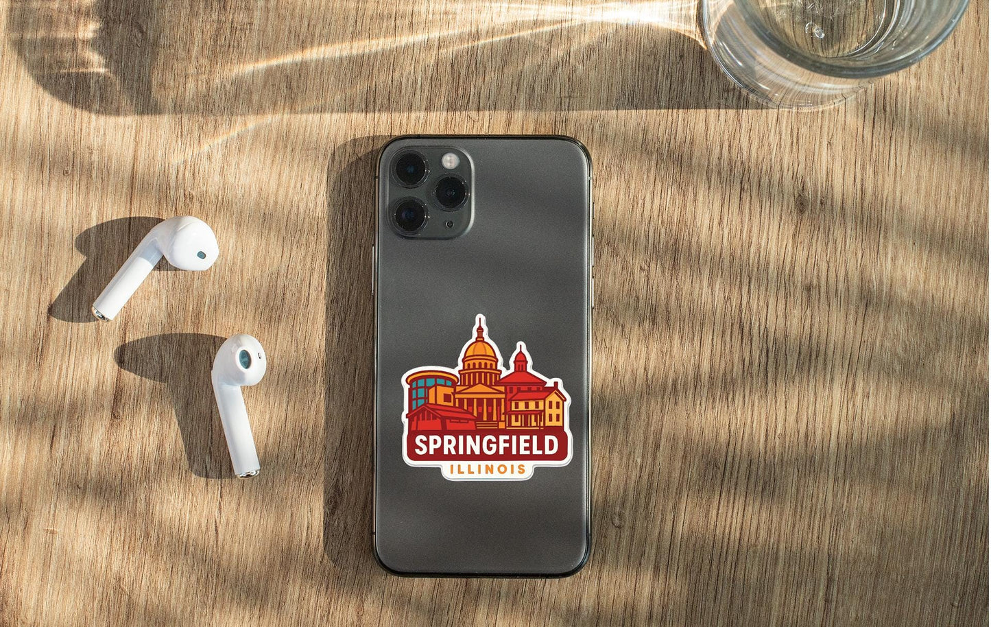 a smartphone with a sticker of the city of Springfield, Illinois, placed on a wooden surface alongside a pair of white earbuds and a glass of water.