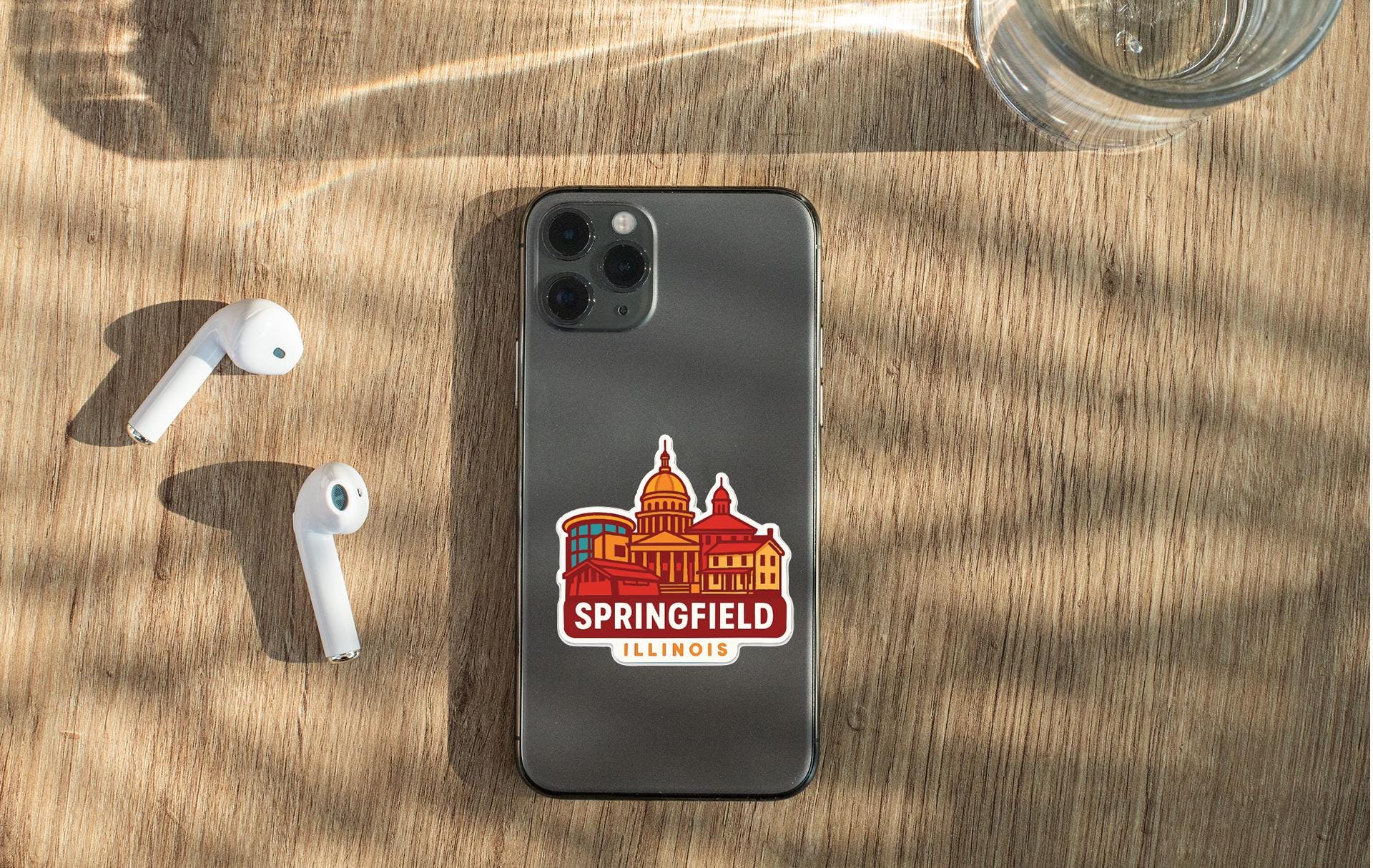 a smartphone with a sticker of the city of Springfield, Illinois, placed on a wooden surface alongside a pair of white earbuds and a glass of water.