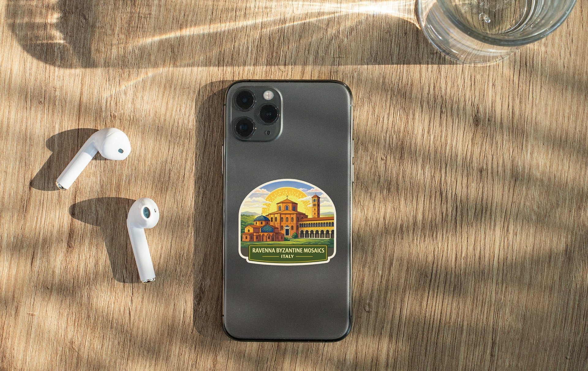 a smartphone with a sticker on it, placed on a wooden surface alongside a pair of white earbuds and a glass of water.