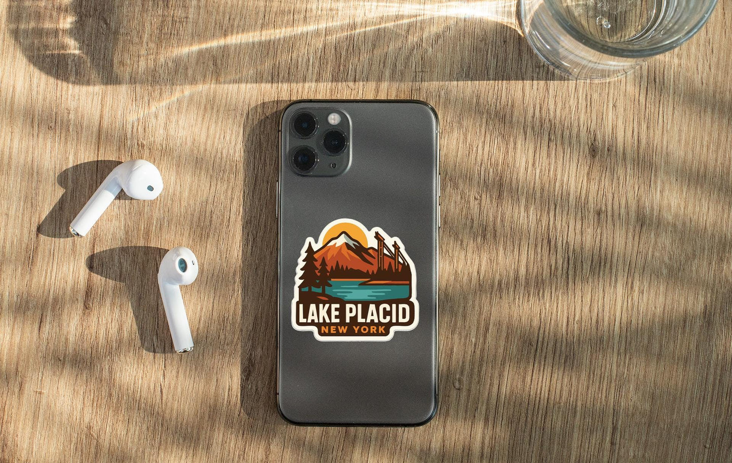 a smartphone with a sticker on the back, placed on a wooden surface alongside a pair of white earbuds and a glass of water.