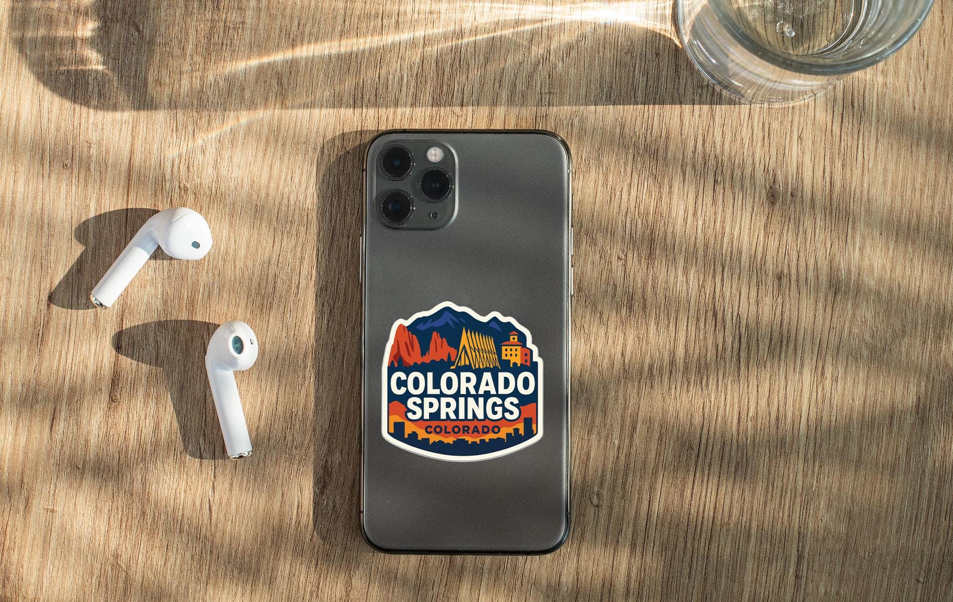 a smartphone with a Colorado Springs, Colorado sticker on its back, along with a pair of white earbuds and a glass of water on a wooden surface.