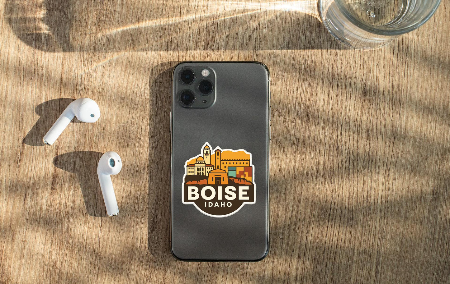 a smartphone with a sticker of Boise, Idaho, on its back cover, along with a pair of white earbuds and a glass of water on a wooden surface.
