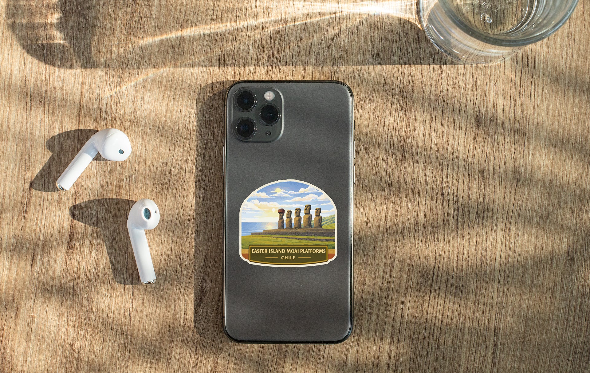 a smartphone with a sticker of Easter Island's Moai statues, a pair of white earbuds, and a glass of water on a wooden surface.