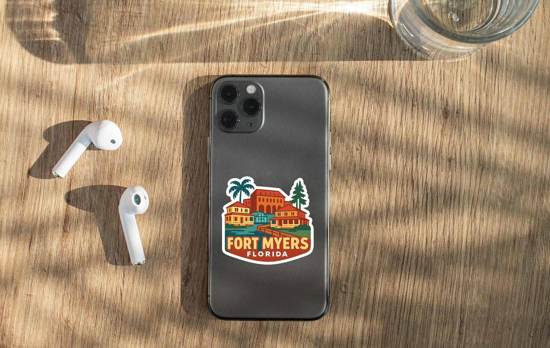 a smartphone with a sticker on the back, placed on a wooden surface alongside a pair of white earbuds and a glass of water.