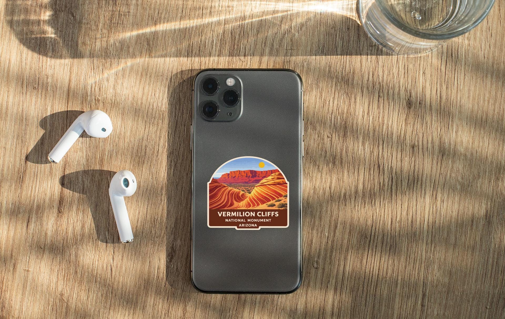 a smartphone with a sticker on it, placed on a wooden surface alongside a pair of white earbuds and a glass of water.