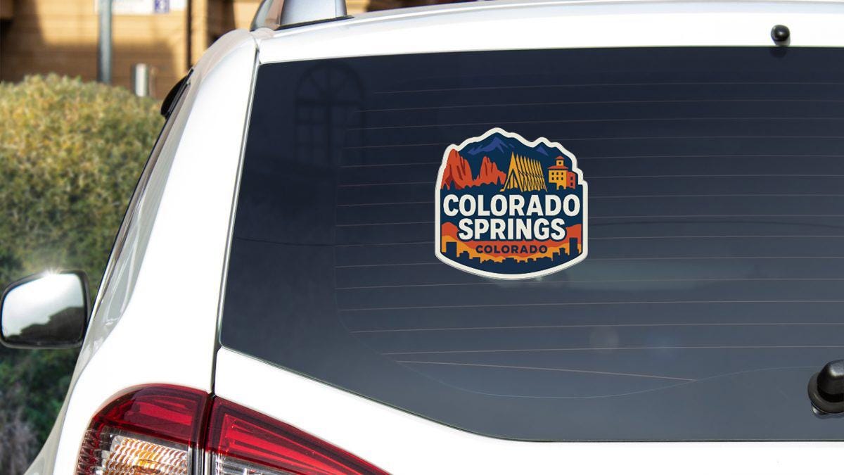 A sticker on the rear window of a white vehicle, featuring the text "COLORADO SPRINGS, COLORADO" along with an illustration of mountains and a building.