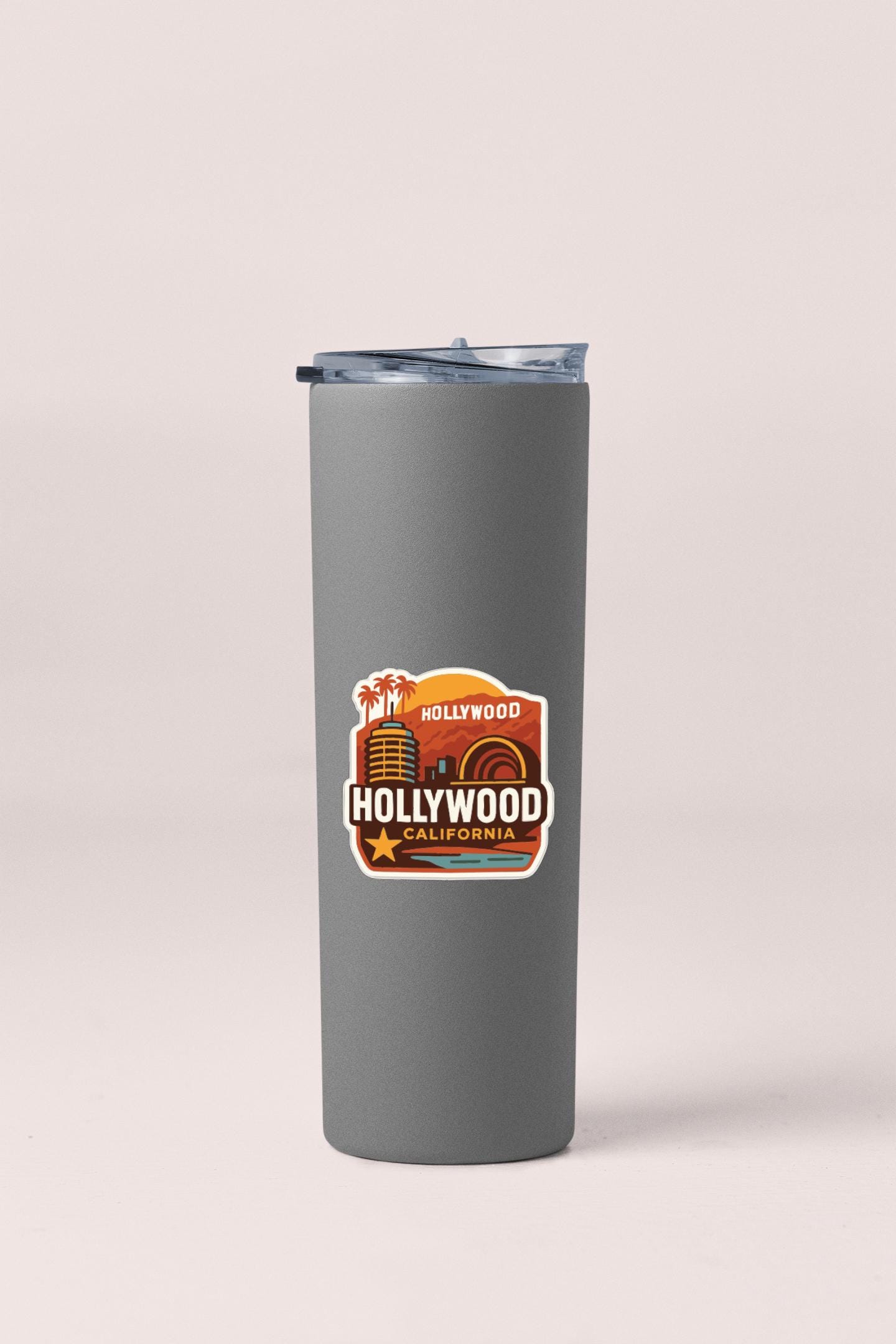 A gray insulated tumbler with a lid and a sticker featuring the text "Hollywood California" and an illustration of a city skyline.