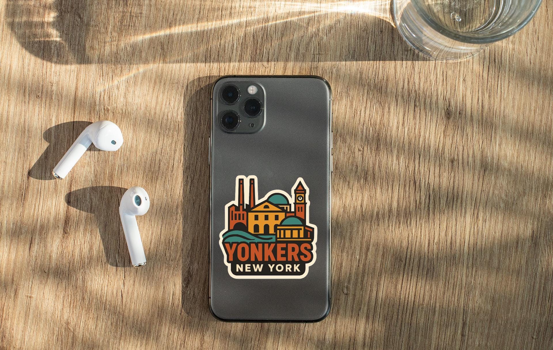 a smartphone with a sticker of the city of New York on it, along with a pair of white earbuds and a glass of water on a wooden surface.