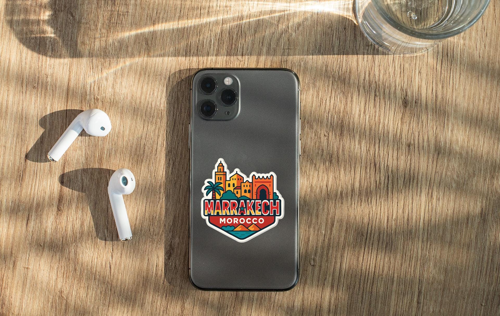 a smartphone with a sticker on the back, placed on a wooden surface alongside a pair of white earbuds and a glass of water.