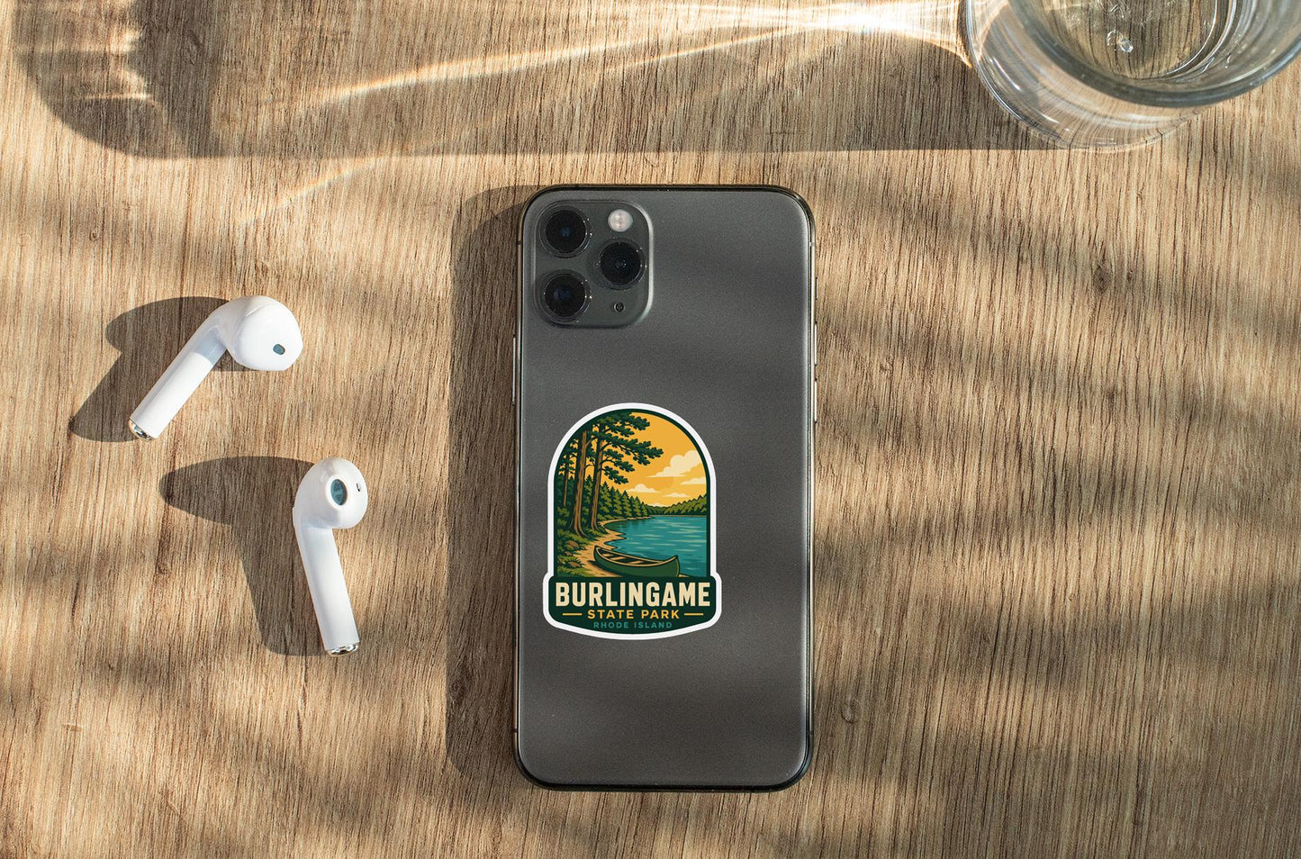 A smartphone with a sticker of Burlingame State Park on its back, along with a pair of white earbuds and a glass of water on a wooden surface.