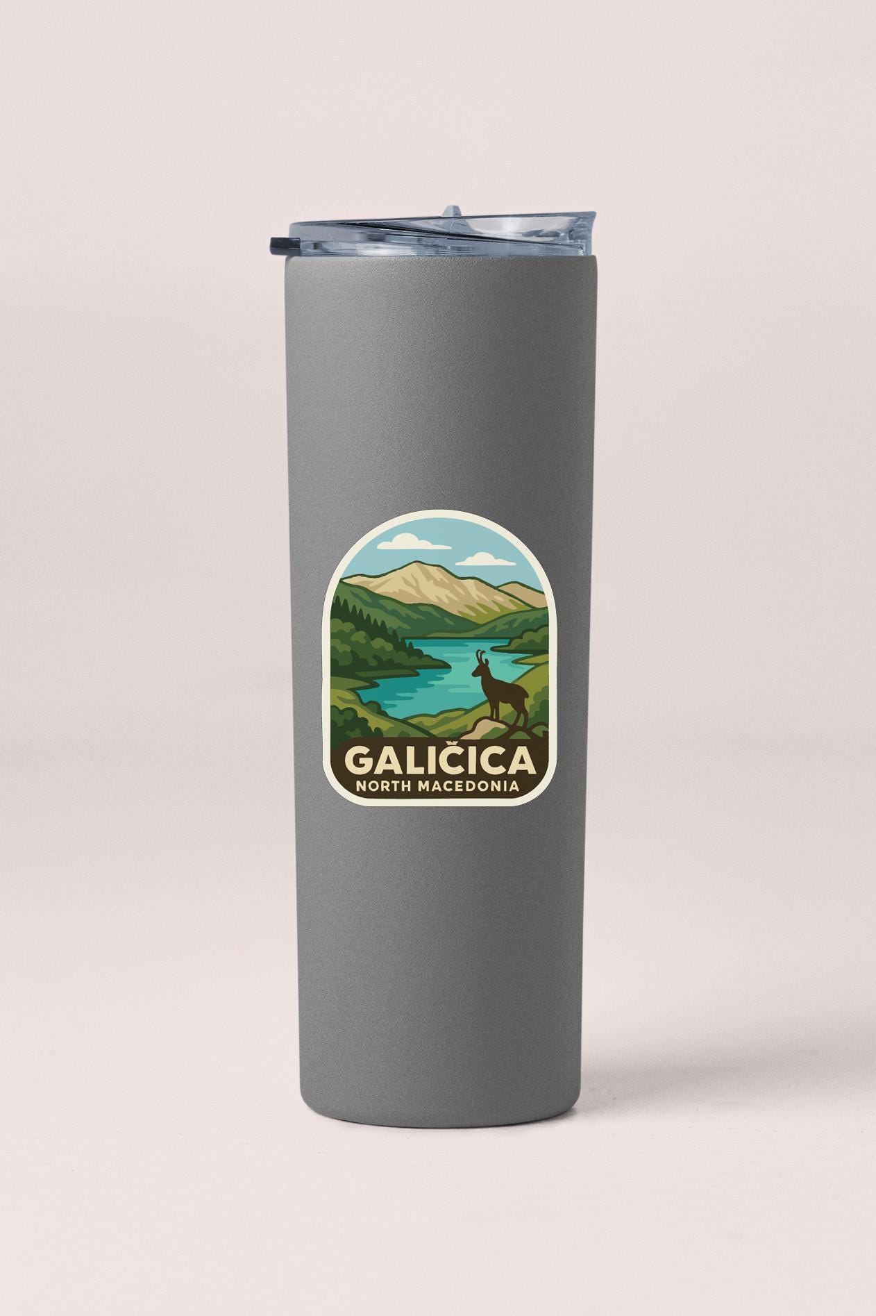 A gray tumbler with a sticker depicting a scenic landscape featuring mountains, a lake, and a deer.