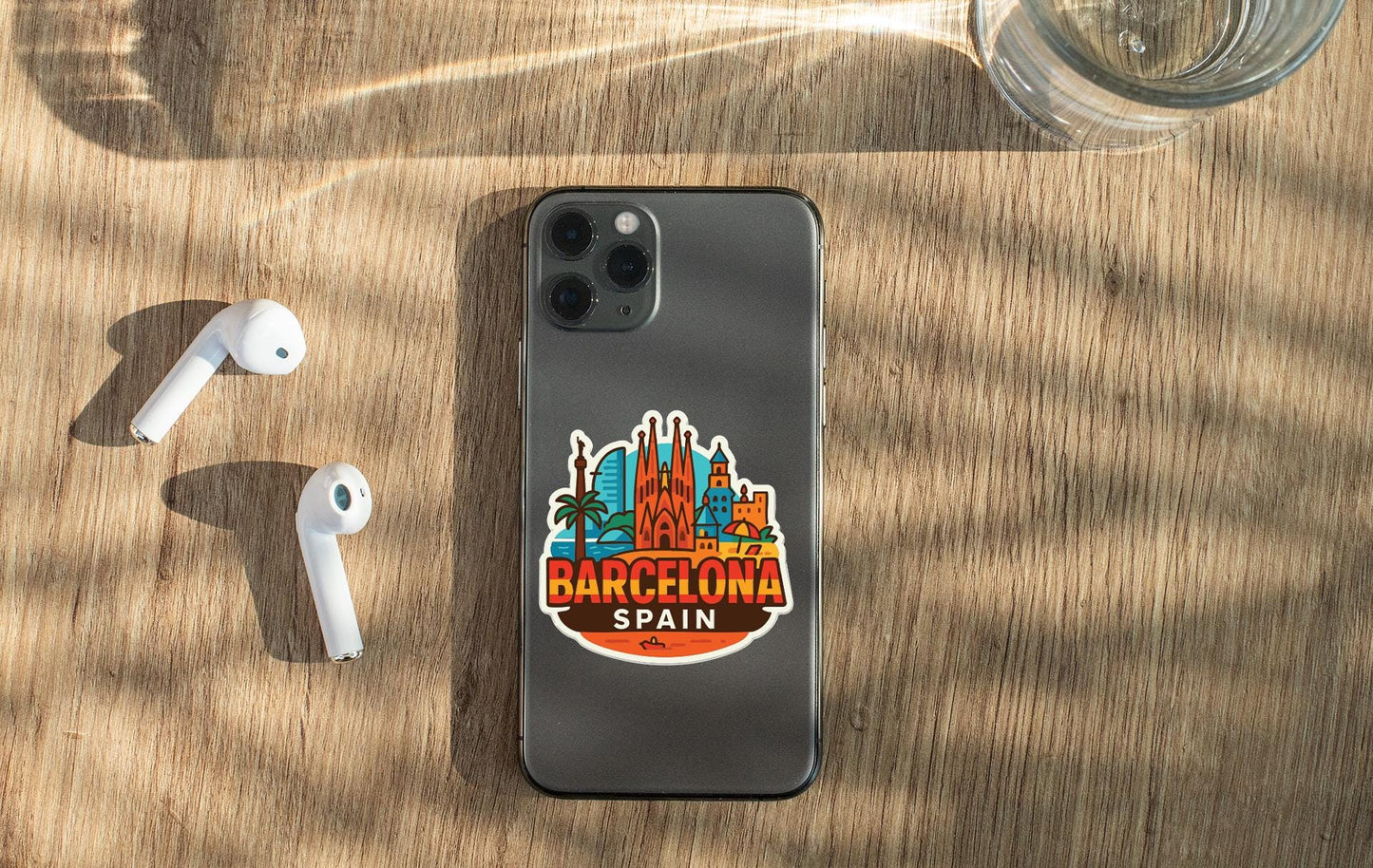 a smartphone with a Barcelona Spain sticker on its back, placed on a wooden surface alongside a pair of white earbuds and a glass of water.