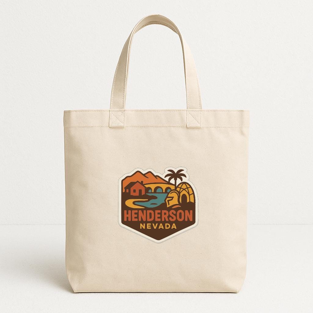 A beige tote bag with a colorful graphic patch on the front, depicting a desert landscape with mountains, palm trees, and a body of water.