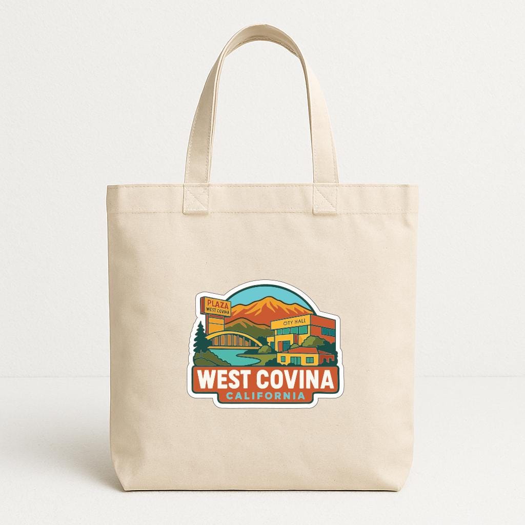 A beige tote bag with a colorful graphic depicting West Covina, California, featuring a mountain, a building, and a sign that says "WEST COVINA CALIFORNIA".