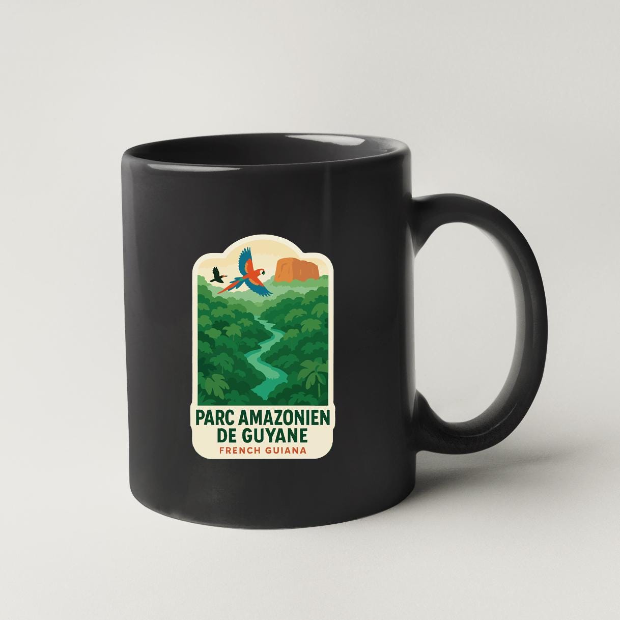 A black coffee mug with a label that says "PARC AMAZONIEN DE GUYANA FRENCH GUYANA" and an illustration of a tropical forest scene with a bird flying over the trees.