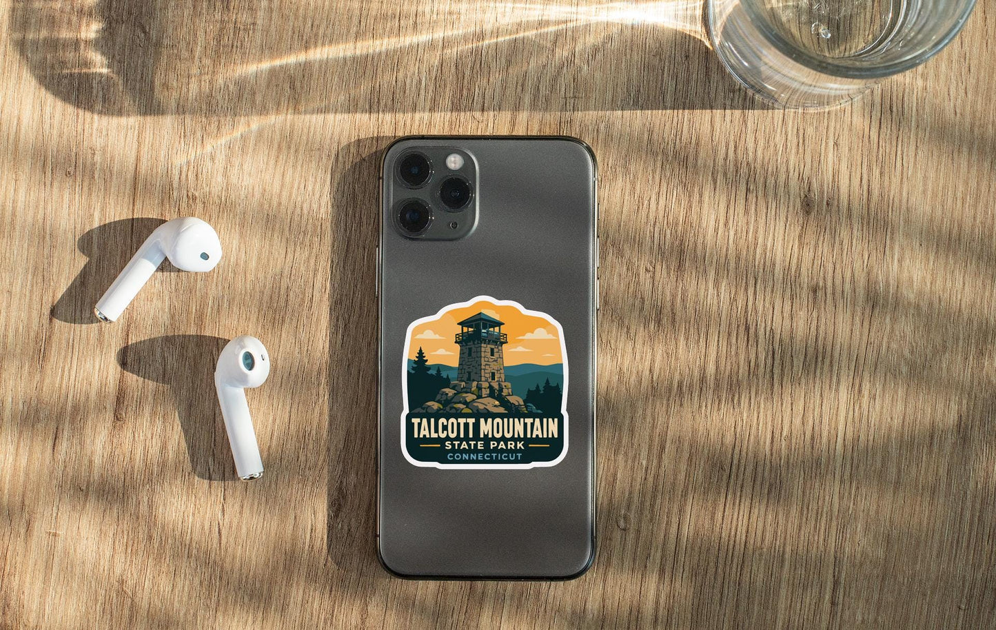 a smartphone with a sticker of Talcott Mountain State Park on its back, along with a pair of white earbuds and a glass of water on a wooden surface.