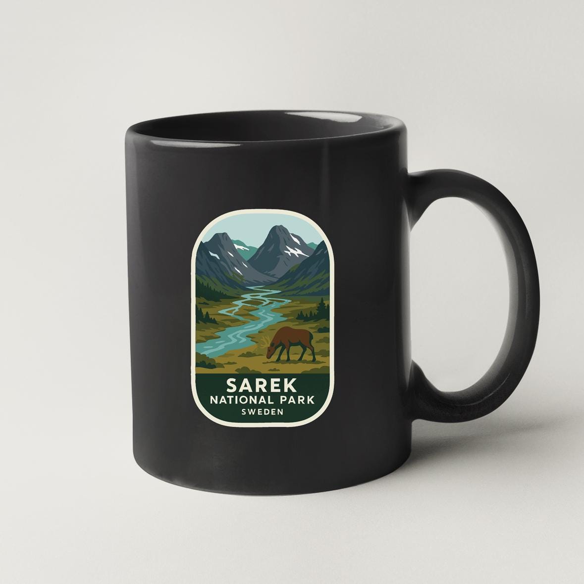 A black coffee mug with a label that features a scenic mountain landscape with a river and a brown animal, likely a bear, in the foreground.