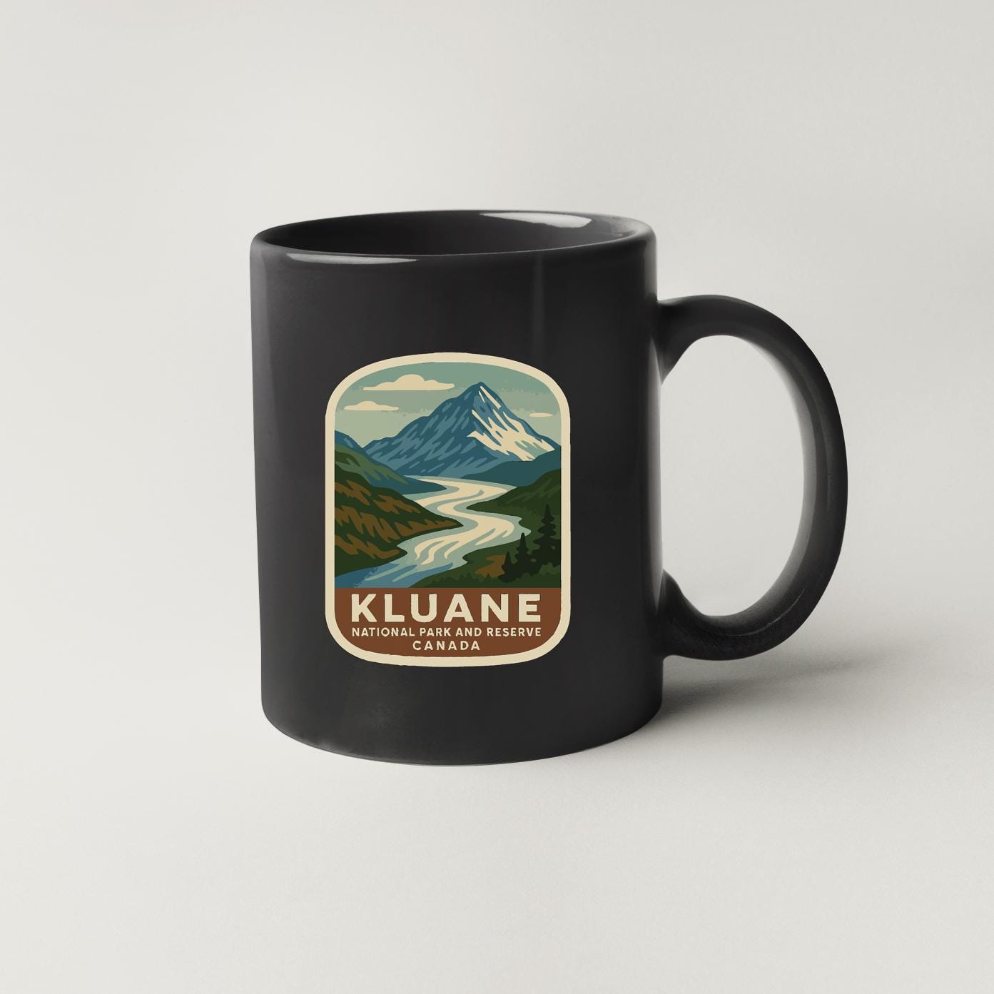 A black coffee mug with a vintage-style illustration of a mountain landscape and a river, along with the text "Kluane National Park and Reserve, Canada".
