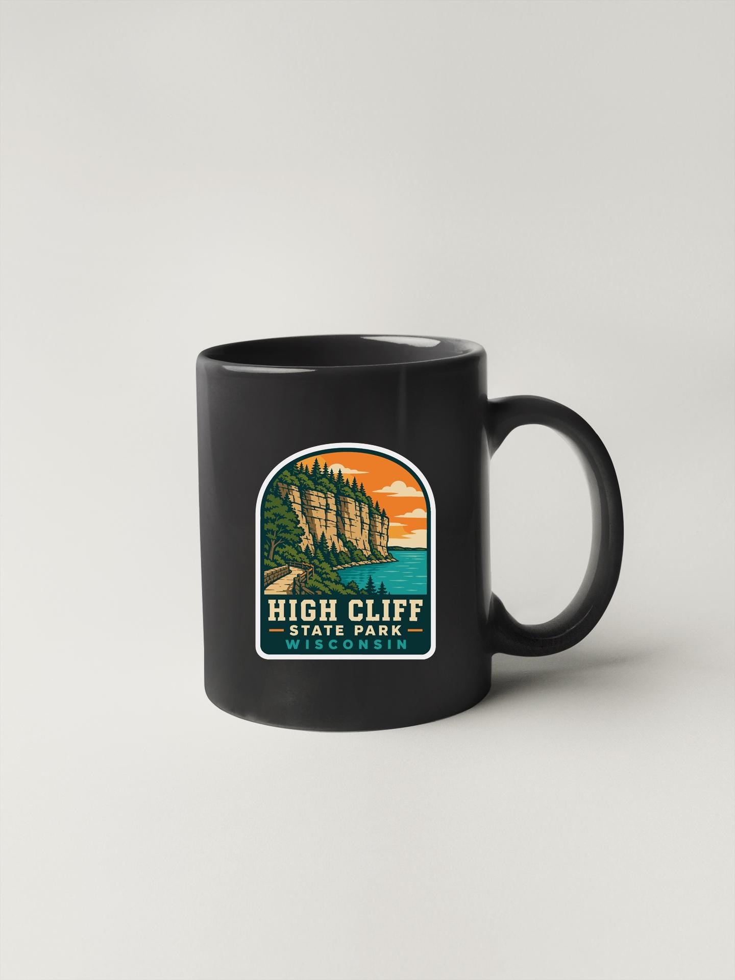 A black coffee mug with a colorful graphic depicting a scenic landscape, including a cliff, trees, and a body of water.