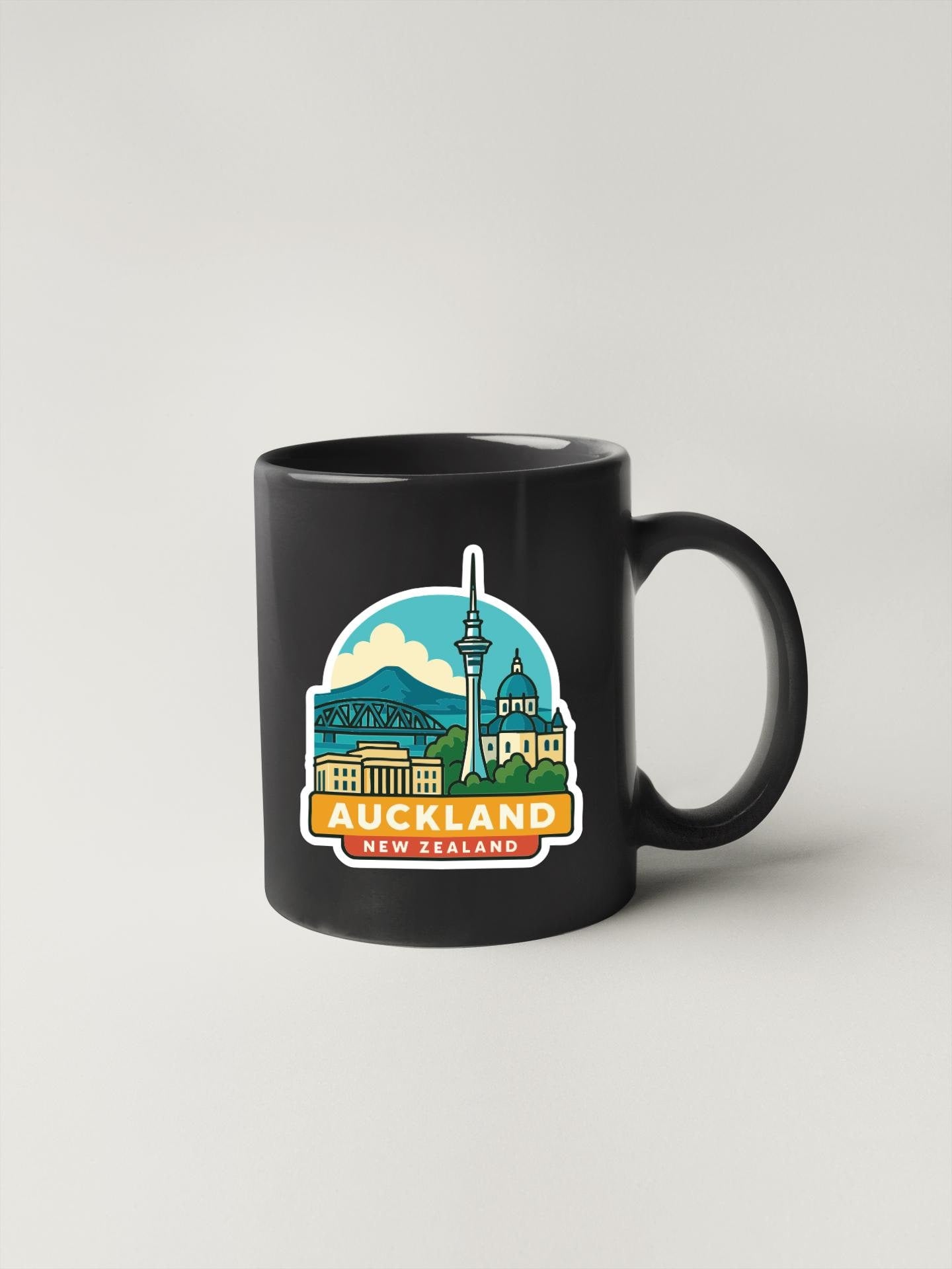 A black coffee mug with a colorful illustration of the city of Auckland, New Zealand, featuring landmarks such as the Sky Tower and the Queen Elizabeth II statue.