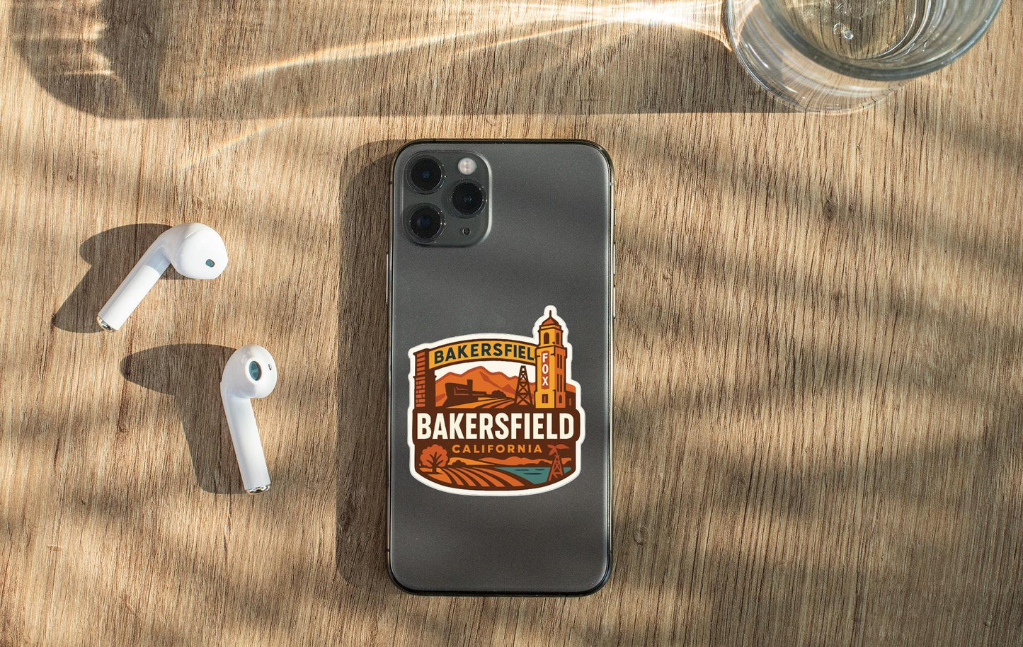 a smartphone with a sticker on the back, a pair of white earbuds, and a glass of water on a wooden surface.