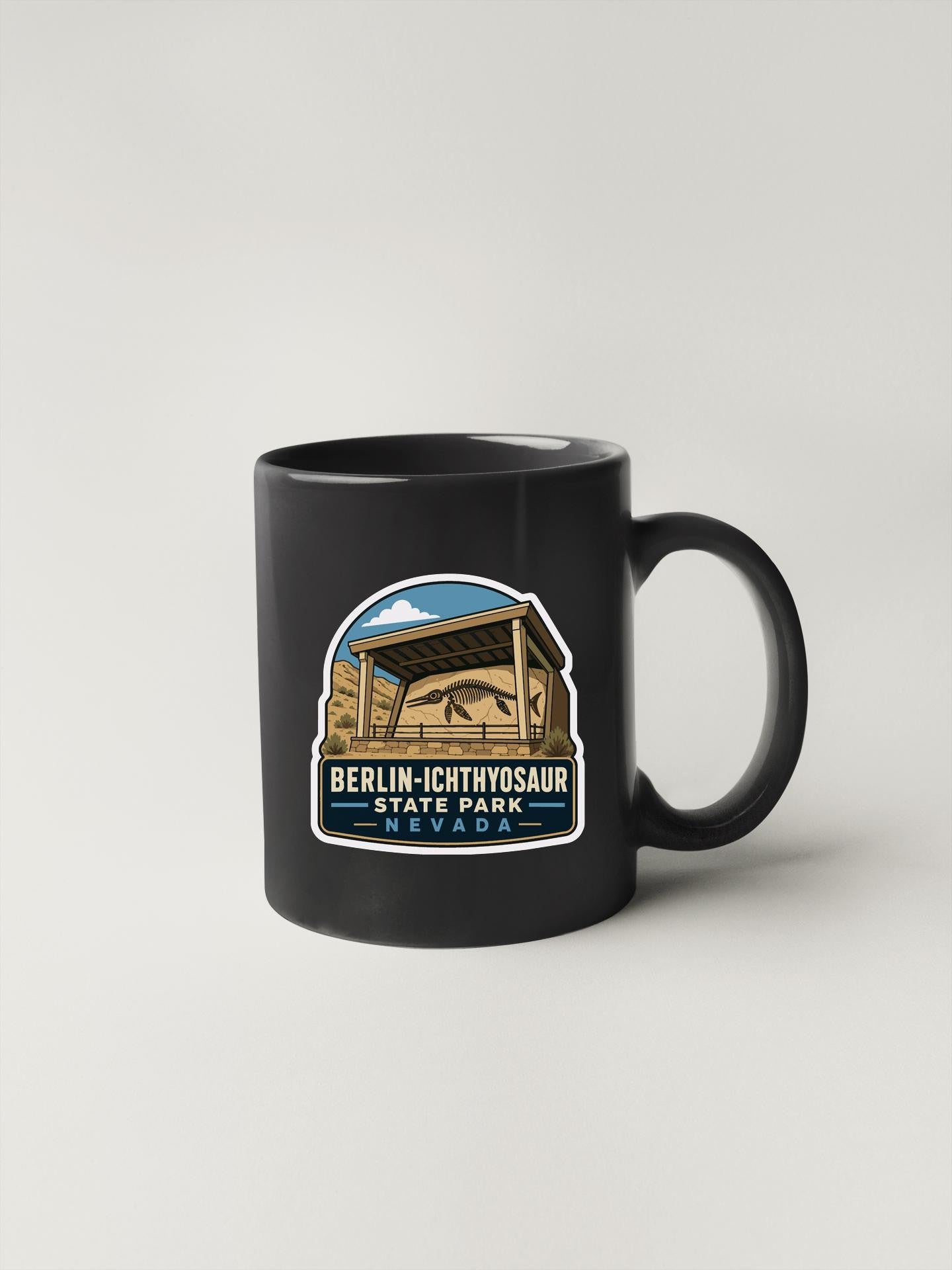 A black coffee mug with a graphic design depicting a building and a dinosaur, along with the text "BERLIN-ICTHYOSAUER STATE PARK NEVADA".