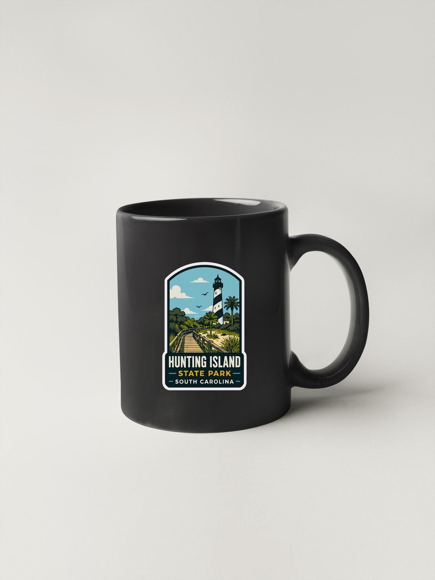 A black coffee mug with a label that says "HUNTING ISLAND STATE PARK SOUTH CAROLINA" and features an illustration of a lighthouse and a road.